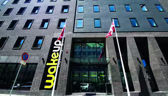 Eksteriør av en moderne murbygning med rektangulære vinduer. Et stort gult og svart "Wakeup"-skilt vises nær inngangen, flankert av to flaggstenger med røde og hvite flagg. Himmelen er klar og blå.