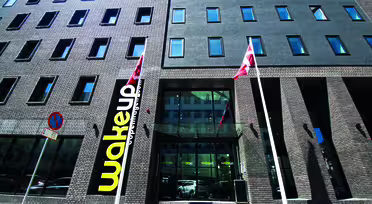 Eksteriør av en moderne murbygning med rektangulære vinduer. Et stort gult og svart "Wakeup"-skilt vises nær inngangen, flankert av to flaggstenger med røde og hvite flagg. Himmelen er klar og blå.