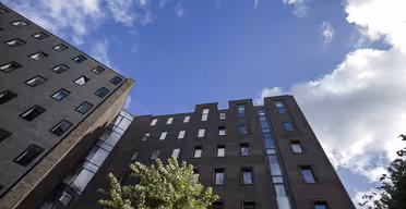 Utsikt over to moderne murbygninger under en blå himmel med spredte skyer. Et tre med grønne blader er i forgrunnen, og bygningenes vinduer reflekterer himmelen.