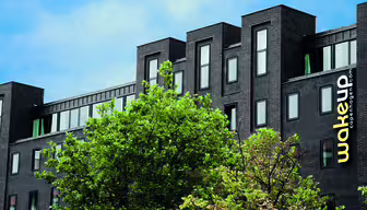 Moderne, mørk mursteinsbygning med store vinduer under en blå himmel. Et knallgult "wakeup copenhagen"-skilt er synlig på siden. Grønne trær skjuler delvis nedre del av bygget.