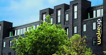 Moderne, mørk mursteinsbygning med store vinduer under en blå himmel. Et knallgult "wakeup copenhagen"-skilt er synlig på siden. Grønne trær skjuler delvis nedre del av bygget.