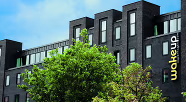 Moderne, mørk mursteinsbygning med store vinduer under en blå himmel. Et knallgult "wakeup copenhagen"-skilt er synlig på siden. Grønne trær skjuler delvis nedre del av bygget.