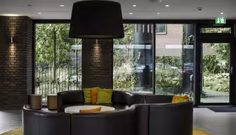 Et moderne salongområde med en sirkulær svart skinnsofa rundt et rundt bord, toppet med fargerike puter. En stor svart lampe henger over. Utenfor store vinduer står grønne trær og en murbygning. Et utgangsskilt er synlig.