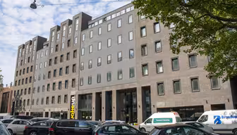 En moderne bygning i flere etasjer med en blanding av murstein og betongfasade, med store vinduer og en fremtredende inngang. Flere biler og varebiler står parkert på gaten foran, med trær som delvis rammer inn scenen.