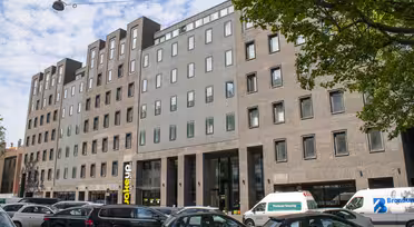 En moderne bygning i flere etasjer med en blanding av murstein og betongfasade, med store vinduer og en fremtredende inngang. Flere biler og varebiler står parkert på gaten foran, med trær som delvis rammer inn scenen.
