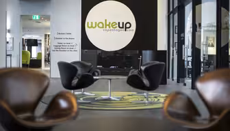 Moderne hotellobby med elegante, brune stoler arrangert rundt små bord. En stor svart sirkel med «wakeup copenhagen.com» er på veggen. Det er et grønt teppe under stolene og naturlig lys fra store vinduer lyser opp rommet.