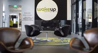 Moderne hotellobby med elegante, brune stoler arrangert rundt små bord. En stor svart sirkel med «wakeup copenhagen.com» er på veggen. Det er et grønt teppe under stolene og naturlig lys fra store vinduer lyser opp rommet.