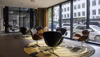 Romslig moderne lobby med svingstoler på mønstrede tepper, store vinduer med utsikt over en bygate. Sykler og parkerte biler synlig utenfor. Naturlig lys fyller rommet og fremhever elegante designelementer.