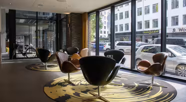 Romslig moderne lobby med svingstoler på mønstrede tepper, store vinduer med utsikt over en bygate. Sykler og parkerte biler synlig utenfor. Naturlig lys fyller rommet og fremhever elegante designelementer.