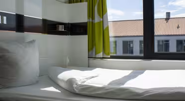 Et lyst hotellrom med en pent oppredd seng, med hvitt sengetøy og en enkelt pute. Grønne og hvite gardiner dekker delvis et stort vindu. Sollys strømmer inn og lyser opp rommet. Utenfor er det utsikt over bygninger med røde tegltak.