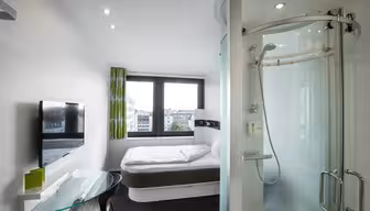 Et moderne hotellrom med en seng ved et stort vindu med utsikt over byen, en montert flatskjerm-TV, et skrivebord i glass og en unik sylindrisk dusj med buet skyvedør. Innredningen er minimalistisk med lyse aksenter.
