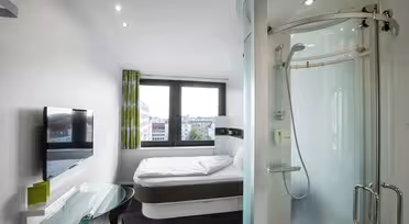 Et moderne hotellrom med en seng ved et stort vindu med utsikt over byen, en montert flatskjerm-TV, et skrivebord i glass og en unik sylindrisk dusj med buet skyvedør. Innredningen er minimalistisk med lyse aksenter.