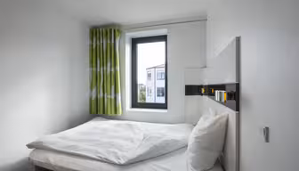 Minimalistisk soverom med en pent oppredd seng, hvitt sengetøy og et vindu med grønne mønstrede gardiner. Vinduet gir utsikt over bygninger utenfor. Rommet har hvite vegger og en moderne, enkel design.