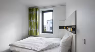 Minimalistisk soverom med en pent oppredd seng, hvitt sengetøy og et vindu med grønne mønstrede gardiner. Vinduet gir utsikt over bygninger utenfor. Rommet har hvite vegger og en moderne, enkel design.