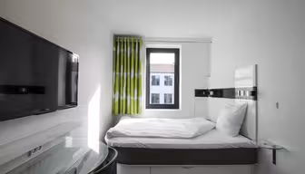 Lite moderne hotellrom med en pent oppredd seng, flatskjerm-TV på veggen og et vindu med grønne og hvite gardiner. Skrivebord og stol med glassplate er i nærheten av sengen. Sollyset strømmer gjennom vinduet.