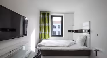 Lite moderne hotellrom med en pent oppredd seng, flatskjerm-TV på veggen og et vindu med grønne og hvite gardiner. Skrivebord og stol med glassplate er i nærheten av sengen. Sollyset strømmer gjennom vinduet.