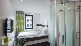Moderne hotellrom med kompakt planløsning. Den har en seng med hvitt sengetøy, et lite rundt skrivebord i glass, en veggmontert flatskjerm-TV og et buet dusjhjørne i glass. Et vindu med grønne og hvite gardiner gir naturlig lys.