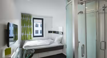 Moderne hotellrom med kompakt planløsning. Den har en seng med hvitt sengetøy, et lite rundt skrivebord i glass, en veggmontert flatskjerm-TV og et buet dusjhjørne i glass. Et vindu med grønne og hvite gardiner gir naturlig lys.