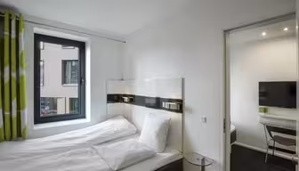 Et moderne hotellrom med dobbeltseng med hvitt sengetøy og hodegjerde i mørkt tre. Et vindu med grønne gardiner er til venstre. Et lite glassbord står ved siden av sengen, og en døråpning fører til et rom med TV og skrivebord.