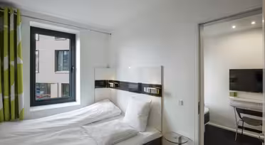 Et moderne hotellrom med dobbeltseng med hvitt sengetøy og hodegjerde i mørkt tre. Et vindu med grønne gardiner er til venstre. Et lite glassbord står ved siden av sengen, og en døråpning fører til et rom med TV og skrivebord.