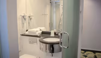 Et moderne bad med vask og benkeplate, pent stablede hvite håndklær og doble veggmonterte hårfønere. Et dusjkabinett i glass og mønstrede gulvfliser er synlige, med grønne og hvite aksenter som gir et friskt preg.