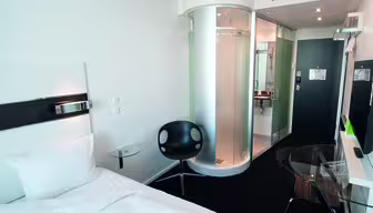 Moderne hotellrom med enkeltseng, hvite vegger og sirkulær dusjkabinett i glass. Det er en svart stol, en veggmontert TV og et skrivebord med glassplate med en brun stol. Inngangsdøren er synlig i bakgrunnen.