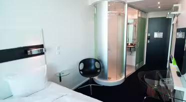 Moderne hotellrom med enkeltseng, hvite vegger og sirkulær dusjkabinett i glass. Det er en svart stol, en veggmontert TV og et skrivebord med glassplate med en brun stol. Inngangsdøren er synlig i bakgrunnen.