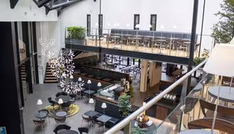 Moderne spisestue med åpen planløsning med to plan. Det nederste nivået har runde bord, stoler og dekorative planter. Det øverste nivået har en rad med bord og stoler. Et stort, kunstnerisk lysarmatur henger i taket, og naturlig lys strømmer inn.