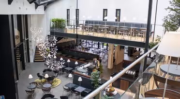 Moderne spisestue med åpen planløsning med to plan. Det nederste nivået har runde bord, stoler og dekorative planter. Det øverste nivået har en rad med bord og stoler. Et stort, kunstnerisk lysarmatur henger i taket, og naturlig lys strømmer inn.