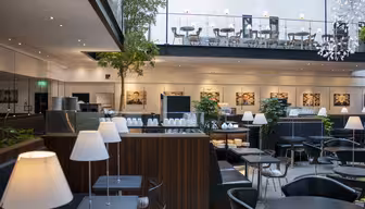 Moderne kaféinteriør med svarte bord og stoler, hvite lamper og pottetrær. En øvre balkong har ekstra sitteplasser. Stor veggkunst og dempet belysning skaper en stilig atmosfære.