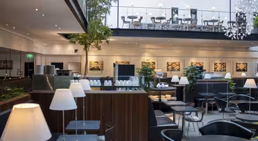 Moderne kaféinteriør med svarte bord og stoler, hvite lamper og pottetrær. En øvre balkong har ekstra sitteplasser. Stor veggkunst og dempet belysning skaper en stilig atmosfære.