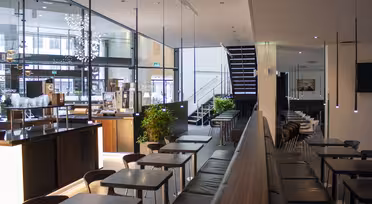 Moderne kaféinteriør med trebord og trestoler, lange sittebenker og pendellys. Store vinduer til venstre gir naturlig lys, og en trapp er synlig i bakgrunnen. Minimalistisk innredning med noen få potteplanter gjennomgående.