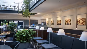 Moderne, romslig kaféinteriør med svarte bord og stoler, hvite hengende lamper og grønne planter. En balkong med ekstra sitteplasser er synlig ovenfor. Veggene er utsmykket med innrammet kunst, og et høyt tre gir et preg av natur.