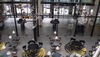 En moderne kaféutsikt ovenfra, med runde bord og stoler på abstrakte gule og sorte mønstrede tepper. Store vinduer slipper inn naturlig lys, og dekorative hengende lys gir eleganse til det romslige interiøret.