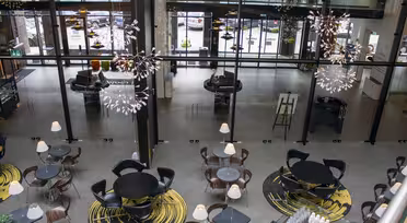 En moderne kaféutsikt ovenfra, med runde bord og stoler på abstrakte gule og sorte mønstrede tepper. Store vinduer slipper inn naturlig lys, og dekorative hengende lys gir eleganse til det romslige interiøret.