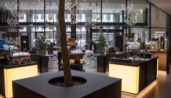 Stilig hotellobby med moderne belysning og innredning. Et stort pottetre i en firkantet plantekasse sitter i midten, omgitt av opplyste disker. Glassvegger avslører en utendørs utsikt. Stoler og bord er arrangert rundt i plassen.