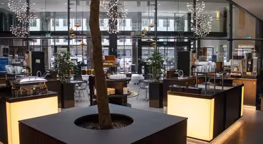 Stilig hotellobby med moderne belysning og innredning. Et stort pottetre i en firkantet plantekasse sitter i midten, omgitt av opplyste disker. Glassvegger avslører en utendørs utsikt. Stoler og bord er arrangert rundt i plassen.