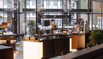 Moderne, godt opplyst bufféområde med elegante trebenker og glassvegger. Stabler av hvite tallerkener og en kaffemaskin er synlige. Dekorativ belysning og frodige grønne planter forsterker den moderne atmosfæren.