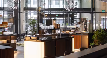 Moderne, godt opplyst bufféområde med elegante trebenker og glassvegger. Stabler av hvite tallerkener og en kaffemaskin er synlige. Dekorativ belysning og frodige grønne planter forsterker den moderne atmosfæren.