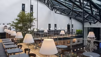 Romslig moderne kafé med glasstak. Har svarte bord med hvite lamper i en sittegruppe med flere nivåer. Naturlig lys lyser opp rommet. Grønne planter gir et snev av natur til den elegante, industrielle designen.