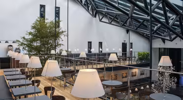 Romslig moderne kafé med glasstak. Har svarte bord med hvite lamper i en sittegruppe med flere nivåer. Naturlig lys lyser opp rommet. Grønne planter gir et snev av natur til den elegante, industrielle designen.