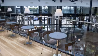 En moderne kafé i en øvre etasje med trebord og trestoler, aksentert av lamper på hvert bord. Designet blander stål og glass, og gir utsikt over den travle gaten nedenfor gjennom store vinduer.
