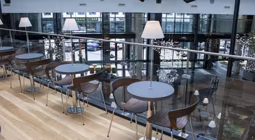 En moderne kafé i en øvre etasje med trebord og trestoler, aksentert av lamper på hvert bord. Designet blander stål og glass, og gir utsikt over den travle gaten nedenfor gjennom store vinduer.