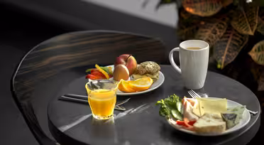 Et rundt bord med frokostpålegg, inkludert en tallerken med frukt og egg, et glass appelsinjuice, et krus kaffe og en tallerken med brød, ost og grønnsaker. En bladplante er i bakgrunnen.