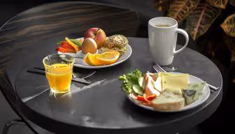 Et lite rundt bord med to tallerkener med frokostmat og drikke. En tallerken har frukt, et egg og en rundstykke; den andre har ost, grønnsaker og brød. Det er et glass appelsinjuice og et hvitt krus kaffe. En plante er i bakgrunnen.