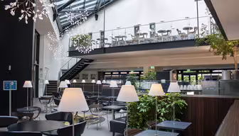 Moderne innendørs kafé med romslig mesanin. Plassen har glassvegger og tak, svarte bord og stoler og hvite hengende lys. En rekke lamper er plassert ved en grønn planteskiller, og skaper en koselig atmosfære.