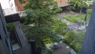 Luftfoto av en murgård med runde bord og stoler som ligger blant frodige grøntområder og små trær. Et treespalier gir delvis skygge. Røde mursteinsbygninger og et metallgjerde omgir det fredelige hageområdet.