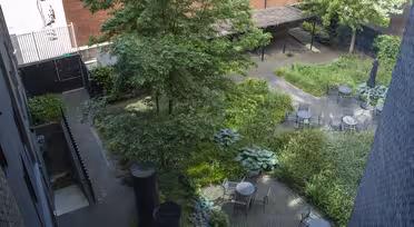 Luftfoto av en murgård med runde bord og stoler som ligger blant frodige grøntområder og små trær. Et treespalier gir delvis skygge. Røde mursteinsbygninger og et metallgjerde omgir det fredelige hageområdet.