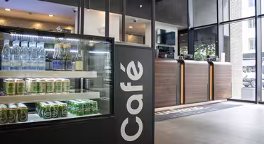 Moderne kaféinteriør med en glassmonter til venstre, som viser flaskevann og hermetikk. Tredisk med utsalgsutstyr i bakgrunnen, nær store vinduer med utsikt til trær og en bygning utenfor.