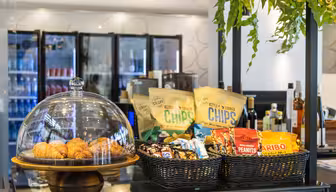 En kafédisk som viser croissanter under en glasskuppel og kurver fylt med diverse snacks, inkludert chips, peanøtter og gummigodteri. Kjøleskap med drikkevarer er synlige i bakgrunnen, sammen med hengende grøntområder over.